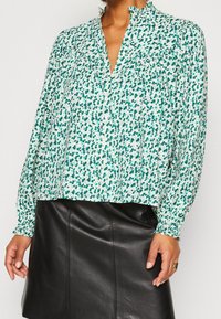 Blumenbluse mit grün-weißem Muster, mit einem Kragen und langen, elastischen Ärmeln. Getragen mit einem schwarzen Lederrock.