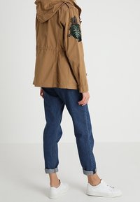 Brown lightweight jacket with a hood and embroidered green leaf design on the sleeve, paired with rolled-up dark blue jeans and white sneakers.