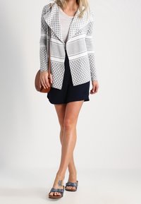 Szary i biały kardigan w wzory z głębokim kołnierzem, połączony z jasną bluzką i granatowymi szortami; noszony z niebieskimi sandałami. Brązowa torba na ramię.