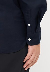 Navy blue shirt with long sleeves, featuring button cuffs and a smooth, textured fabric. Visible white buttons on cuffs.