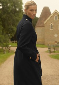 Žena s plavom kosom u tamnom dugom kaputu, stoji na šljunčanom putu u vrtu s zelenim drvećem i kamenim zdanjem u pozadini.