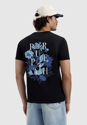 Man draagt een zwart T-shirt met een blauw bloemenpatroon en witte letters "PURPLE PATH" op de achterkant, lichte spijkerbroek en een witte honkbalpet.