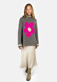 Woman in beige satin skirt, tan boots, and oversized gray sweater with large pink flower and fringed hem, standing on white background.