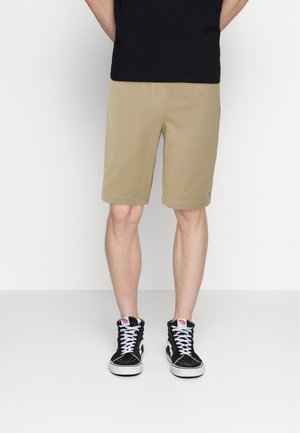Beige bomulds shorts med elastisk talje, der når til midt lår, parret med sorte højtop sneakers med hvide snørebånd og røde detaljer.
