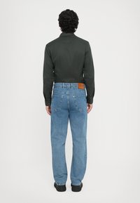 Camisa de manga larga verde oscuro, jeans de denim azul rectos, parche de cuero visible en la cinturilla y zapatos negros.