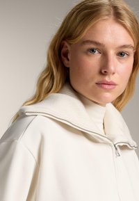 Biela zipová bunda s ribovaným golierom. Mäkká tkanina, uvoľnený strih. Golier je textúrovaný, so strieborným zipsom. Jednoduchý, minimalistický dizajn.