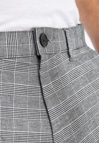 Pantaloni grigi a quadri realizzati in tessuto strutturato, con un bottone nero prominente in vita e motivi a scacchi a contrasto.