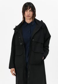 Casaco longo preto feito de um tecido leve, com fecho de correr, dois bolsos frontais e capuz. O design é oversized e estruturado.