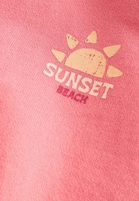 Kahvatu kollase päikese graafika valge "SUNSET" ja roosa "BEACH" teksti kohal tekstuurse roosa kangastel.