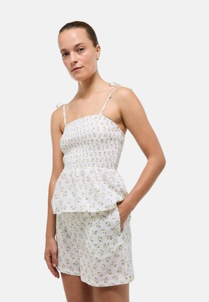 Femme portant un haut sans manches blanc à fleurs avec corsage smocké et short taille haute assorti, debout avec une main dans la poche.