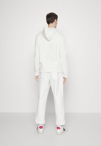 White hoodie with a drawstring, paired with white, rolled-up pants. Sneakers feature grey and navy accents. Minimalist design, smooth texture.