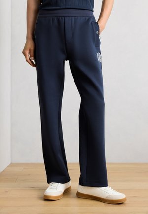 Személy tengerészkék nadrágot visel oldalzsebekkel és logóval, fehér tornacipővel párosítva, fa padlón áll egy sima fal előtt.