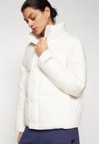 Dikke witte jas met een hoge kraag, voorzien van een logo op de mouw. Glad materiaal en een boxy vorm met een glanzende afwerking.