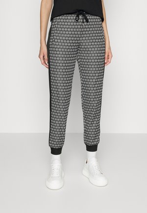 Widok z przodu osoby noszącej czarne dresy z białym powtarzającym się wzorem logo "MK", czarnymi paskami po bokach, czarnymi mankietami, białymi skarpetkami i białymi trampkami.