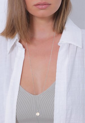 Collier en argent avec un pendentif en perle, porté sur un top gris à côtes. L'arrière-plan est une chemise blanche texturée. Le collier présente des accents délicats.