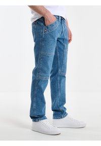 Heledad sinine denim püksid sirge sääre disainiga, mitme taskuga ja kulunud pesuga. Kombineeritud valgete spordijalatsitega.