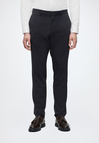 Homme portant un pantalon noir slim fit, une chemise blanche et des mocassins en cuir marron foncé à pampilles, debout devant un fond uni.