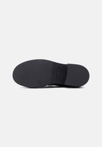 Semelle de chaussure en caoutchouc noir avec une texture lisse et une partie avant arrondie. Elle présente un léger talon et une discrète marque de la marque sur le côté.