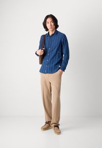 Cămașă albastră cu nasturi, cu un model subtire în dungi, pantalon bej și încălțăminte maro, purtată cu o geantă de umăr maro.