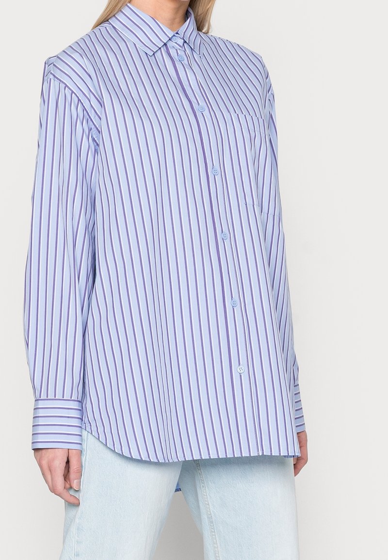Lyseblå skjorte med knapper og lodrette lilla striber, lange ærmer, krave og en brystlomme. Stoffet ser glat og skarpt ud.