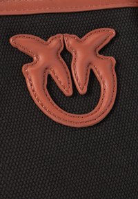 Črn teksturiran material z izstopajočim rjavim usnjenim emblemom v obliki dveh stiliziranih ptic z vidnimi šivi.