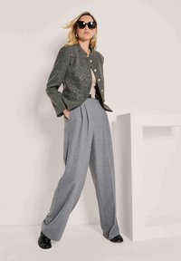 Vrouw in een oversized donkergrijze blazer, geplooide lichtgrijze wijde broek, zwarte riem, zwarte schoenen en grote zwarte zonnebril, poseert.