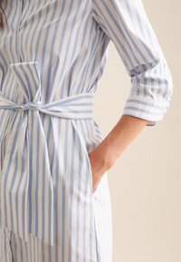 Kvinde iført en lyseblå og hvid vertikalt stribet kjole med 3/4 ærmer og bælte bundet om livet, med den ene hånd i sidelommen.