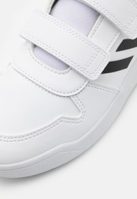 Białe buty sportowe z skórzaną cholewką, siateczkowym językiem i dwoma rzepami. Czarne akcenty po bokach dodają kontrastu. Teksturowana gumowa podeszwa.