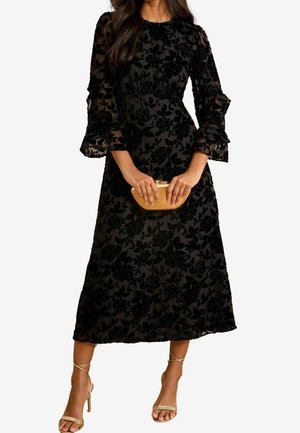 Femme portant une robe midi en velours noire à imprimé floral avec des manches trois-quarts à volants, tenant une pochette beige et portant des talons beige à brides.