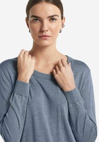 Ljusblå långärmad topp med vid halsringning och åtsittande ärmar, tillverkad av mjukt material. Modellen håller försiktigt tyget nära halsringningen.