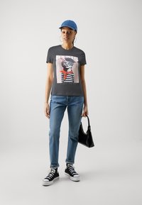 Grå T-skjorte med et kattemotiv som har på seg briller og et rødt skjerf, kombinert med lyseblå jeans og svarte høy-top sneakers. Svart håndveske.