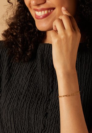 Goldene Kettenarmband am Handgelenk, mit länglichen Gliedern. Strukturierter schwarzer Pullover mit hervortretenden Mustern im Hintergrund.