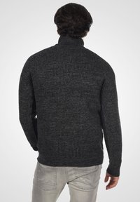 Suéter de punto gris oscuro con cuello alto, patrón texturizado, mangas largas y dobladillo acanalado. Se lleva sobre pantalones gris claro.