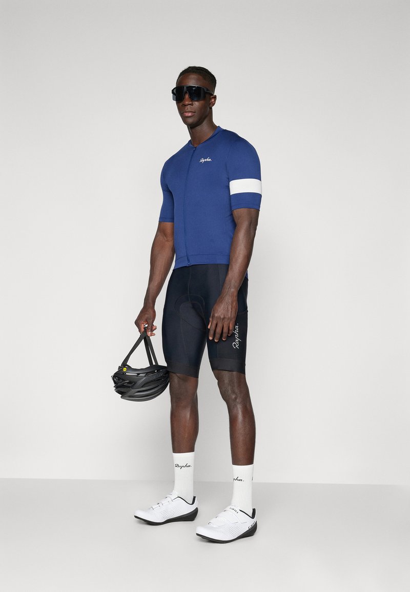 Blå cykeltrøje med en hvid stribe, sorte shorts og hvide cykelsko. Modellen holder en sort hjelm og har solbriller på.