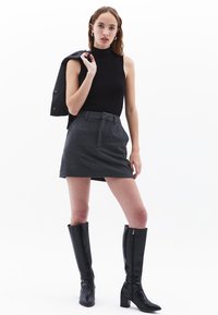 Zwarte mouwloze top, grijze mini-jurk met riem, en kniehoge zwarte leren laarzen. Model houdt een zwarte jas op haar schouder.