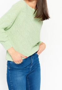 Un pull en tricot vert clair avec une coupe décontractée, un col rond et un motif texturé. Associé à un jean en denim bleu, les mains dans les poches.