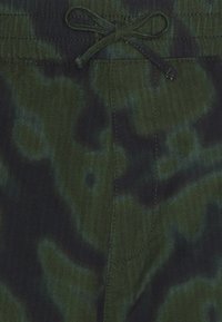 Vihreä, maastokuvioitu kangas, jossa ruudullinen tekstuuri, nyörivyötäröllä ja näkyvillä ompelusaumoilla.