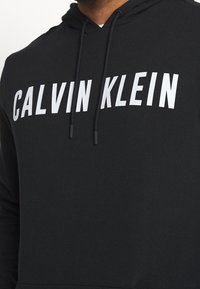 Sudadera negra de tejido suave con un gran logo blanco de "CALVIN KLEIN" en el pecho. Incluye cordones ajustables en la capucha.