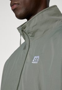 Myk jakke i dempet grønn, med høy krage, glidelås foran, og en liten brodert logo med et geometrisk design.