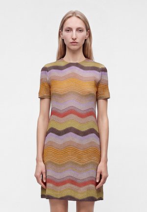 Femme aux longs cheveux blonds portant une robe à manches courtes avec des rayures ondulées horizontales dans des tons violet, orange, marron, rouge et jaune.