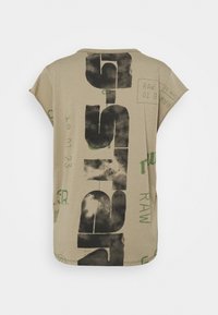 Beige short-sleeve shirt with large distressed black vertical text "RAW" and smaller green text and graphic elements scattered across the back.