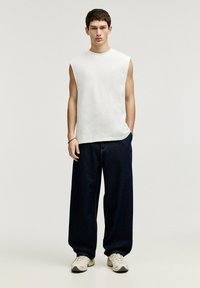 Weißes ärmelloses Top, locker sitzende dunkelblaue Jeans, helle Sneakers. Der Stoff wirkt weich, die Jeans haben eine dezente Steppnaht. Einfaches Design.