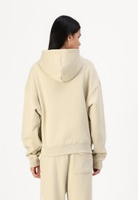 Beige hoodie with a large hood, dropped shoulders, and ribbed cuffs. Features a relaxed fit and a small pocket on the back.