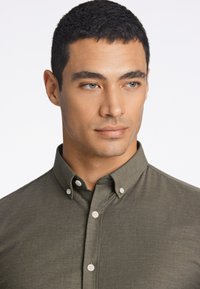 Camisa abotonada verde oliva con cuello, que presenta botones blancos y una tela suave y texturizada. Primer plano de la mitad superior de la camisa.