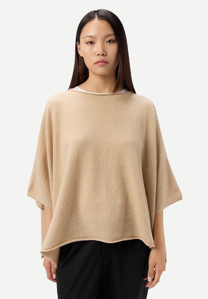 Junge Frau mit langen dunklen Haaren, die einen lockeren beigen Poncho über einem weißen Oberteil und schwarzen Hosen trägt, steht vor einem einfarbigen Hintergrund.