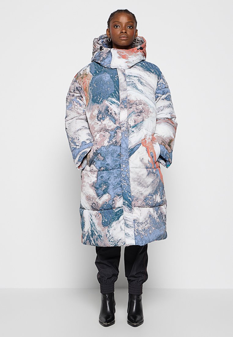 Manteau rembourré avec un motif abstrait bleu, blanc et rose, doté d'un col montant, d'une capuche et de poches avant. Porté par-dessus un pantalon noir et des bottes.