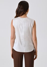 Blusa bianca senza maniche con una texture liscia, orlo arrotondato e dettagli di cucitura centrale, abbinata a pantaloni marroni a vita alta.