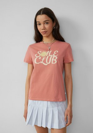 Roze katoenen t-shirt met de print "S A L E C L U B" en een zonafbeelding, gecombineerd met een lichtblauwe geplooide rok van dunne stof.