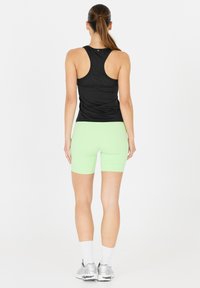 Kvinde med brunt hestehale, iført sort racerback tanktop, lysegrønne cykelshorts, hvide sokker og sølvfarvede løbesko, stående med ryggen til.