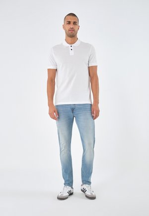White polo shirt with short sleeves and black buttons, paired with light blue jeans and white sneakers with black accents. Simple design.